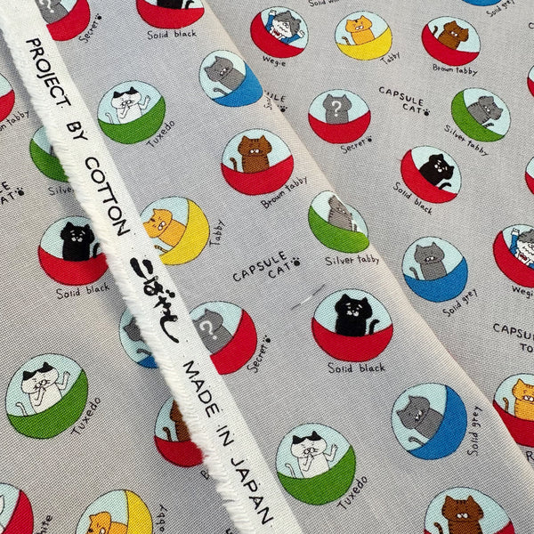 Selvage of Capsule Toy Cats cotton canvas fabric in gray by Cotton Kobayashi featuring playful cats inside capsule toy machines on a gray background
