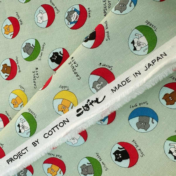 Selvage of Capsule Toy Cats cotton canvas fabric in green by Cotton Kobayashi featuring playful cats inside capsule toy machines on a green background