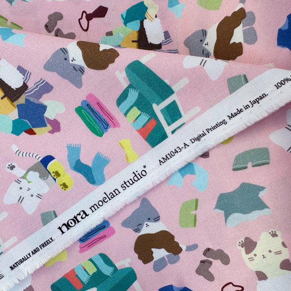 Selvage of Dressed Up Cats cotton fabric in pink by Nora featuring playful cats trying on outfits, surrounded by laundry piles and scattered clothes