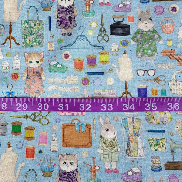 Scale of Dressmaker cotton linen fabric by Yukiemon for Hokkoh featuring delicately illustrated sewing notions, dressmaking tools, vintage sewing machines, and sweet bunny and kitty dressmakers on a blue background, imported from Japan