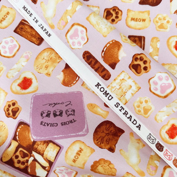 Selvage of Neko Cookie Can cotton Oxford fabric by Komu Strada featuring cat-shaped cookies, biscuits, and cookie tins on a pink background