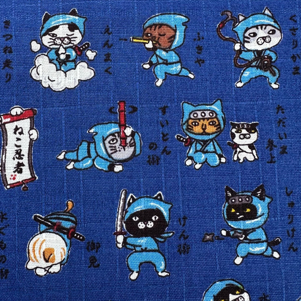 Close up of Neko Ninja blue cotton dobby fabric by Cosmo Textiles, imported from Japan. Features playful ninja cats on a textured cotton fabric.