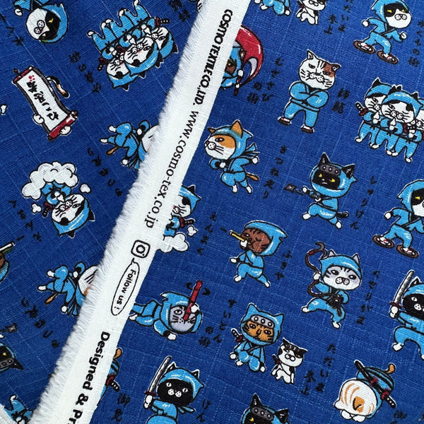 Selvage of Neko Ninja blue cotton dobby fabric by Cosmo Textiles, imported from Japan. Features playful ninja cats on a textured cotton fabric.
